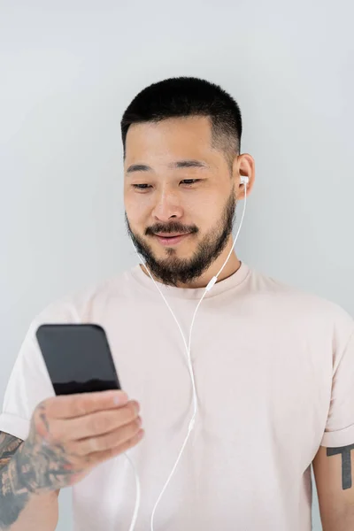 Веселий азіатський чоловік в дротових навушниках слухає музику і тримає смартфон ізольований на сірому — стокове фото