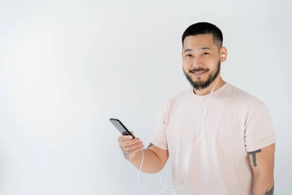 Joyful asian man in wired earphones listening music and holding smartphone isolated on grey — Stock Photo