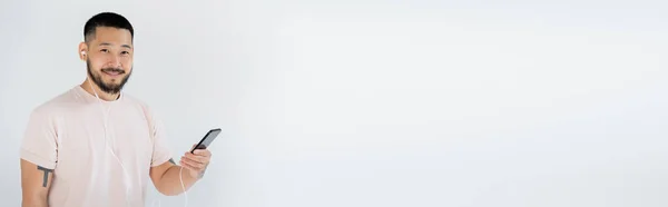 Joyful asian man in wired earphones listening music and holding smartphone isolated on grey, banner — Stock Photo