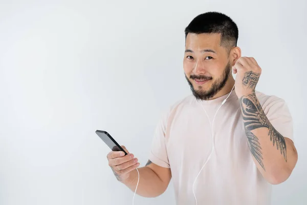 Hombre asiático tatuado con auriculares con cable mientras escucha música y sostiene el teléfono inteligente aislado en gris - foto de stock