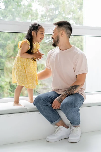 Feliz tatuado hombre sentado en windowsill y mirando alegre asiático hija - foto de stock