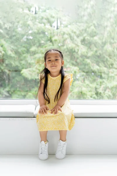 Longitud completa de alegre asiático chica en amarillo vestido sentado en suave almohada en windowsill - foto de stock
