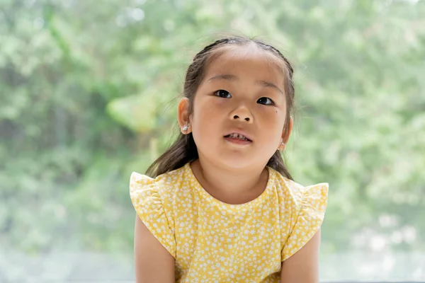 Retrato de morena y preescolar chica asiática en vestido amarillo cerca de la ventana en casa - foto de stock