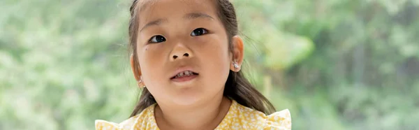 Portrait de fille asiatique brune et préscolaire en robe jaune près de la fenêtre à la maison, bannière — Photo de stock