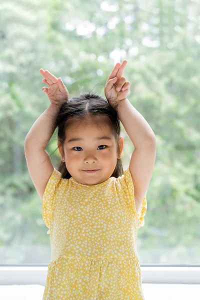 Portrait of brunette and preschooler asian girl in yellow dress showing bunny ears near window at home — Stock Photo