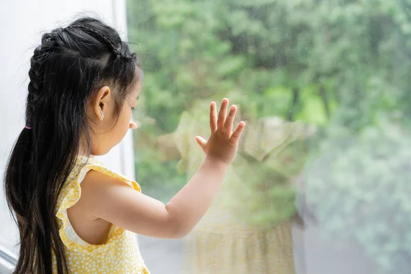 Brunette and preschooler asian girl in yellow dress looking through window at home — Stock Photo