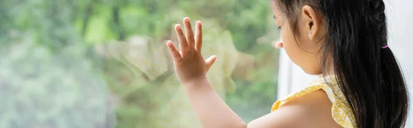 Morena e pré-escolar menina asiática em vestido amarelo olhando através da janela em casa, banner — Fotografia de Stock