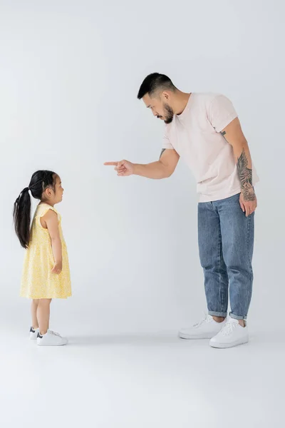 Full length of asian father pointing at preschooler daughter while punishing her on grey — Stock Photo