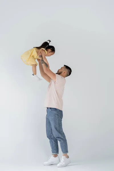 Full length of asian man lifting daughter in yellow dress on grey — Stock Photo