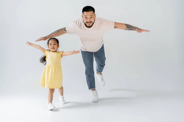 Volle Länge der fröhlichen asiatischen Vater spielen mit Tochter in gelbem Kleid auf grau — Stockfoto