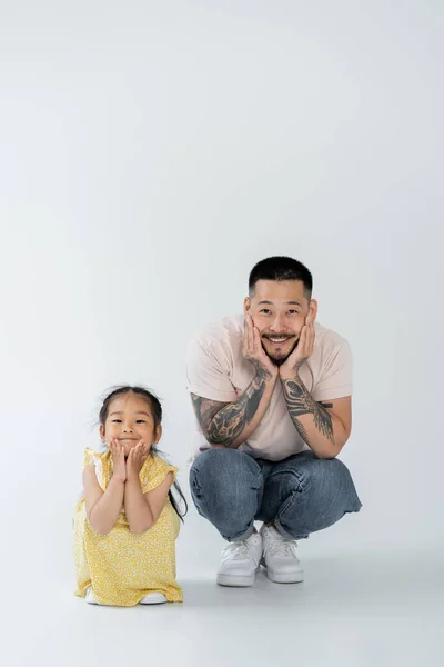 Full length of joyful asian father and cheerful daughter in yellow dress sitting on grey — Stock Photo