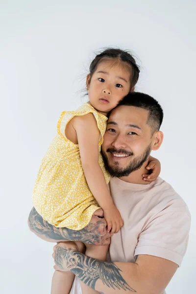 Счастливый азиатский отец с бородой, держа в руках дошкольницу в платье, изолированном на сером — стоковое фото