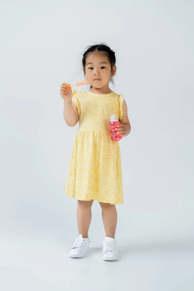 Full length of asian girl in yellow dress holding bottle with soap bubbles on grey — Stock Photo