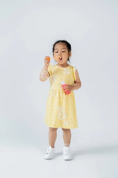 Pleine longueur de l'enfant d'âge préscolaire asiatique en robe jaune tenant bouteille avec bulles de savon sur gris — Photo de stock