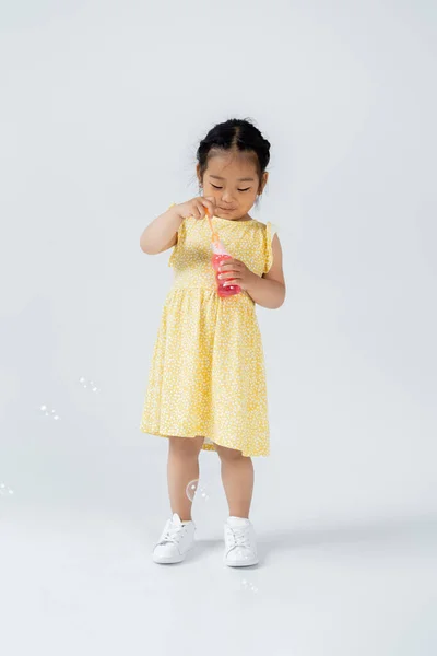 Longitud completa de niño preescolar asiático en vestido amarillo celebración botella con pompas de jabón en gris - foto de stock