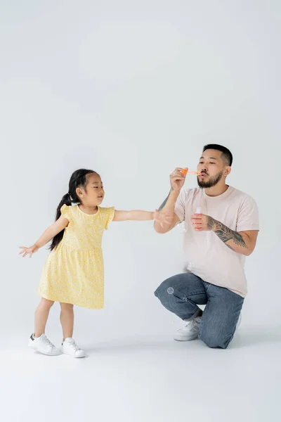 Asian father blowing soap bubbles near cheerful preschooler daughter on grey — Stock Photo