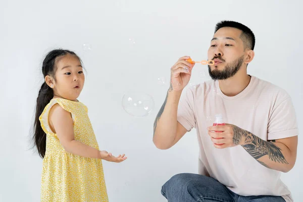 Asiatico padre soffiando sapone bolle vicino sorpreso in età prescolare figlia isolato su grigio — Foto stock