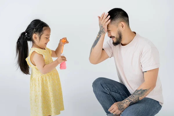 Asian preschooler kid in yellow dress holding bottle with soap bubbles near tattooed father isolated on grey — Stock Photo