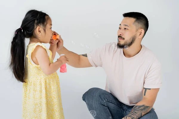 Asian preschooler kid in yellow dress blowing soap bubbles near tattooed father on grey — Stock Photo