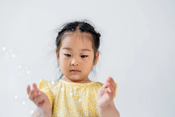 Portrait of asian preschooler girl in yellow dress near blurred soap bubbles isolated on grey — Stock Photo