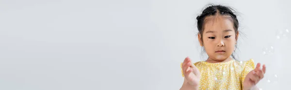 Portrait of asian preschooler girl in yellow dress near soap bubbles isolated on grey, banner — Stock Photo