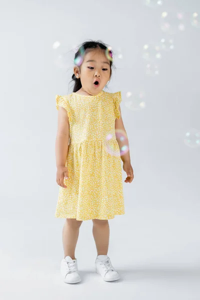 Full length of shocked asian preschooler girl in yellow dress standing soap bubbles on grey — Stock Photo