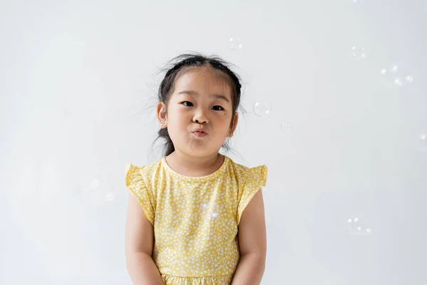 Retrato de asiático preescolar chica en amarillo vestido pucheros labios cerca cerca de jabón burbujas aislado en gris - foto de stock