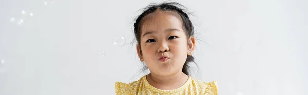 Портрет азиатской дошкольницы, дующейся губами возле мыльных пузырей, изолированных на сером, баннер — стоковое фото