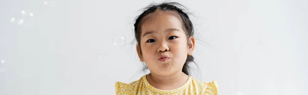 Portrait of asian preschooler girl pouting lips near near soap bubbles isolated on grey, banner — Stock Photo