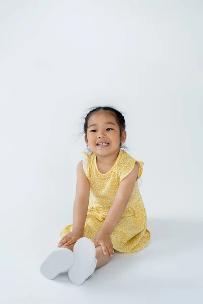 Повна довжина радісної дошкільної дитини в сукні, що сидить на сірому — стокове фото