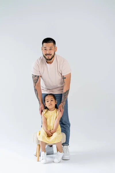 Full length of smiling asian man with tattoos standing near preschooler daughter on grey — Stock Photo