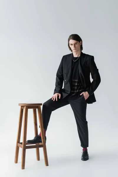 Повна довжина стильного чоловіка в костюмі позує біля стільця на сірому фоні — стокове фото