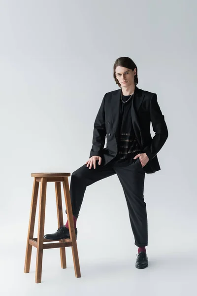 Full length of stylish man in suit posing near chair on grey background — Stock Photo