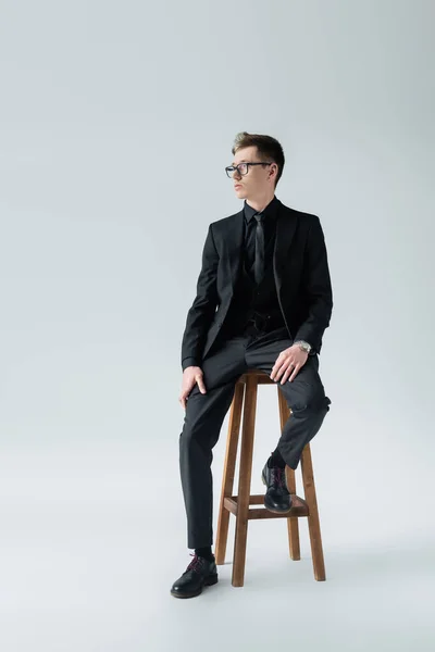 Full length of elegant man in eyeglasses sitting on chair on grey background — Stock Photo