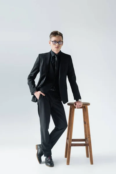 Homem elegante em terno e óculos posando perto da cadeira isolada no cinza — Fotografia de Stock