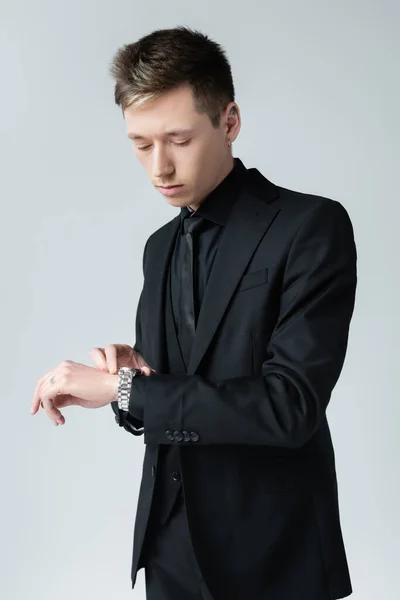 Hombre joven en traje negro mirando reloj de pulsera aislado en gris - foto de stock