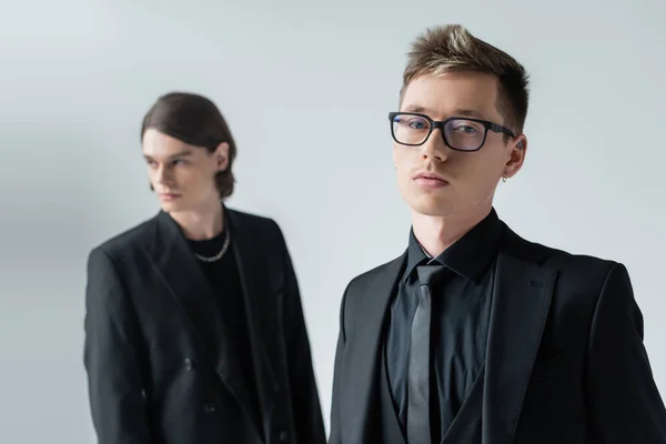 Young man in eyeglasses looking at camera near blurred boyfriend isolated on grey — Stock Photo