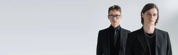 Young gay looking at camera near boyfriend in formal wear and eyeglasses isolated on grey, banner — Stock Photo