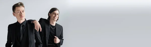 Cheerful gay in formal wear looking at camera near boyfriend isolated on grey, banner — Stock Photo