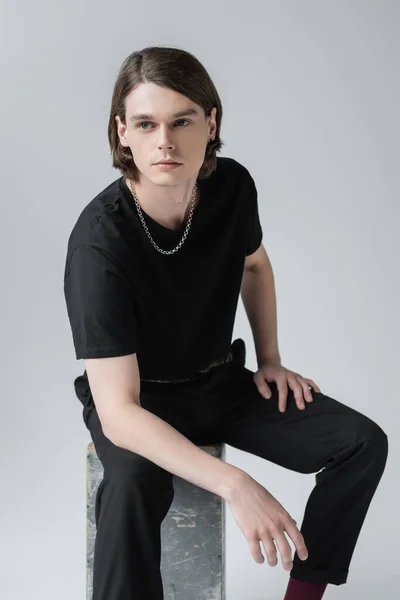 Stilvoller junger Mann schaut weg, während er auf Betonklötzen sitzt, isoliert auf Grau — Stockfoto