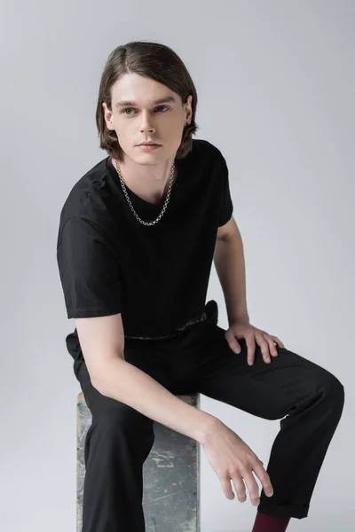 Stylish young man looking away while sitting on concrete block isolated on grey — Stock Photo