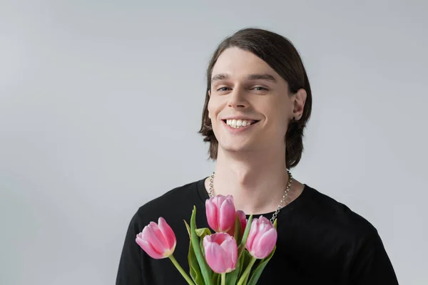 Joven positivo sosteniendo tulipanes rosados aislados en gris - foto de stock