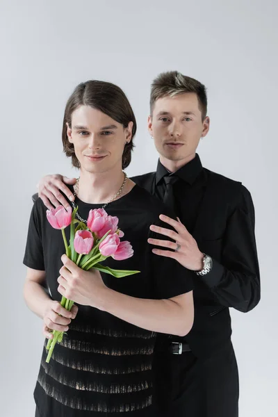 Junge gay umarmen lächelnden Freund mit Tulpen isoliert auf grau — Stockfoto