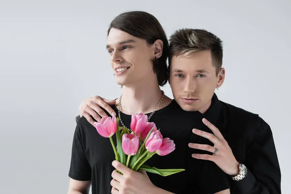 Junge Homosexuell umarmt Freund mit Tulpenblumen isoliert auf grau — Stockfoto