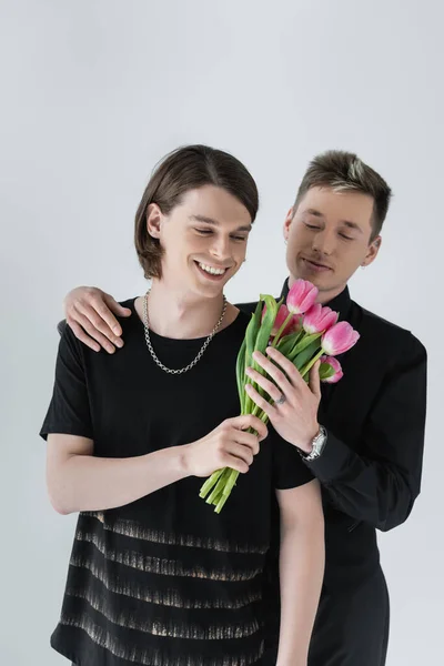 Young gay smiling while holding flowers near boyfriend isolated on grey — Stock Photo