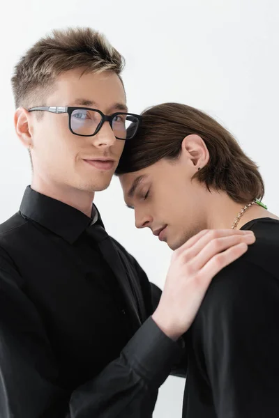 Sonriendo gay en gafas mirando cámara y abrazando novio aislado en gris - foto de stock