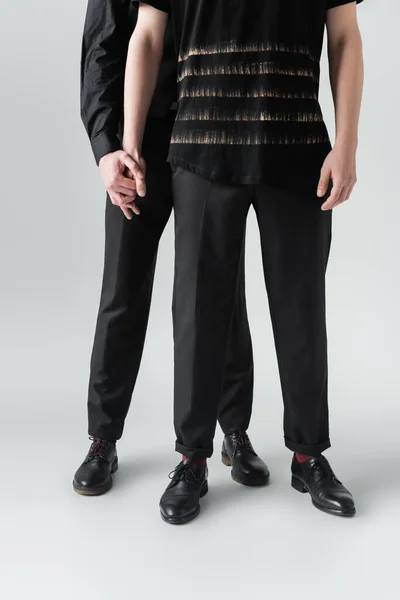 Обрезанный вид гей-пары в обуви и черной одежде, держащихся за руки на сером фоне — стоковое фото