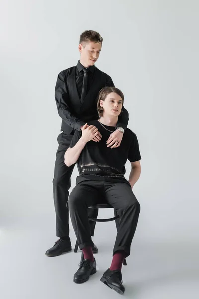 Elegante gay abrazando novio sentado en silla en gris fondo - foto de stock