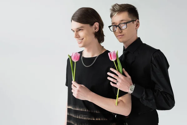 Smiling gay holding pink tulip near boyfriend in eyeglasses isolated on grey — Stock Photo