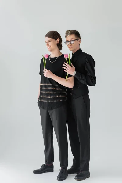 Повна довжина стильної пари гей, що тримає рожеві тюльпани на сірому фоні — стокове фото